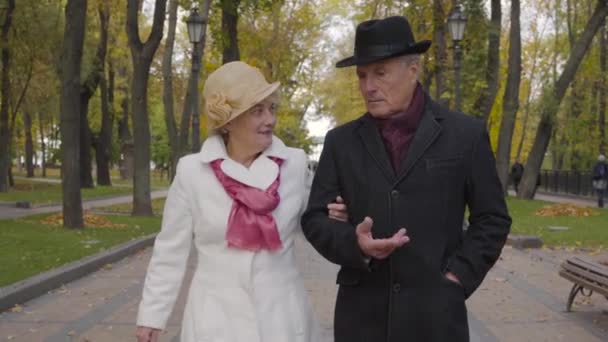 Close-up of a senior Caucasian couple walking in the autumn park. Mature man in black coat and hat talking to his adorable wife and smiling. — ストック動画