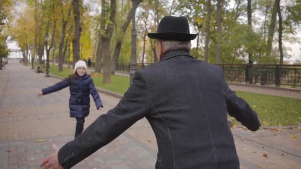 Zarif siyah şapkası ve paltosu olan yaşlı beyaz bir adamın torununun yukarı koşup onu büyütmesini ve kucaklamasını beklemesi. Gülümseyen kız büyükbabasına koşuyor.. — Stok video