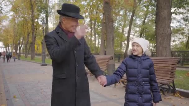 Hombre mayor caucásico con elegante sombrero negro y abrigo paseando con su nieta por el callejón en el parque de otoño. Niña linda hablando con su sabio abuelo y sonriendo . — Vídeos de Stock