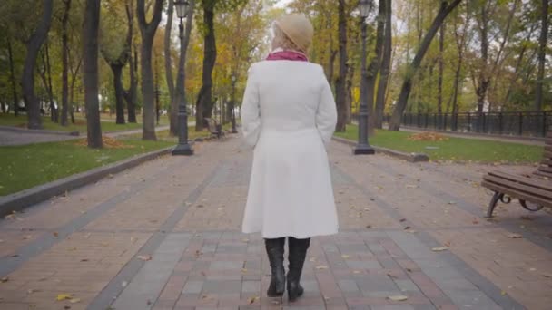 Vista posteriore della donna caucasica in elegante cappello cloche beige e cappotto bianco passeggiando lungo il vicolo. Maturo signora riposo dopo il suo pensionamento nel parco autunnale . — Video Stock