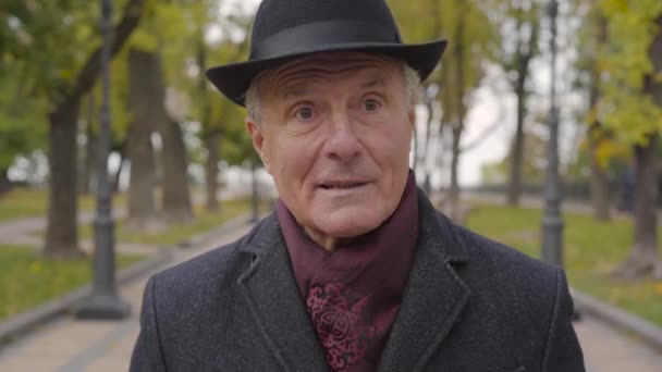 Close-up of a mature Caucasian man with hazel eyes dressed in elegant black hat and coat standing in the autumn park and talking. Wise mister explaining something. — Stock Video