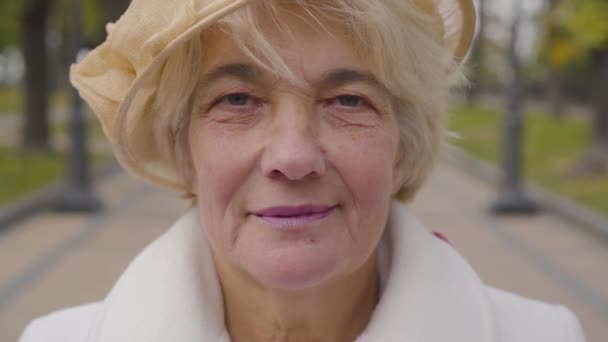 Primer plano extremo de la mujer blanca mayor con ojos azules mirando a la cámara. Señora madura en sombrero de cloche beige de pie en el parque de otoño . — Vídeo de stock