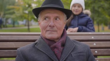 Arka plandan gelen neşeli beyaz kız bankta oturan zarif, klasik elbiseli olgun bir adamın gözlerini kapıyor. Torunu, güzel dedesine sonbahar parkında sürpriz yapıyor.