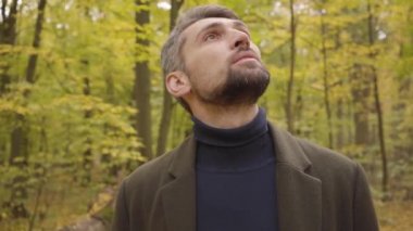 Ormanda dikilip yukarı bakan yakışıklı, beyaz bir adamın yakın çekimi. Sakin bir sonbahar gününün tadını çıkaran sıradan elbiseli bir adam..
