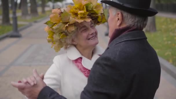 Glimlachende volwassen blanke vrouw in een krans van gele bladeren die danst met haar lieve man in het steegje in het herfstpark. Positieve senior familie dating buiten. — Stockvideo