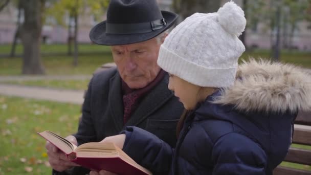 Бічний погляд на дорослого кавказького чоловіка в класичному одязі, який сидить на лавці зі своєю внучкою і читає книгу в червоній обкладинці. Досить усміхнена дівчина уважно слухає свого мудрого діда.. — стокове відео