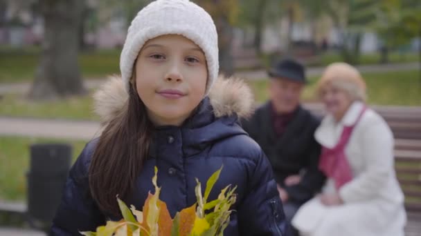 Leende vit tjej i vit hatt som håller ett gäng gula blad och tittar på kameran. Hennes morförälder sitter på bänken i bakgrunden. — Stockvideo