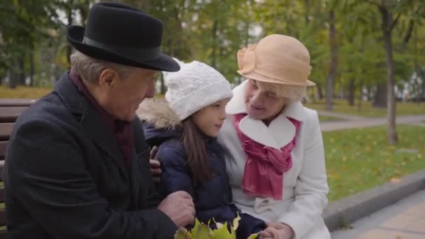 Ein älteres kaukasisches Paar sitzt mit seiner Enkelin auf der Bank im herbstlichen Park. Großeltern in eleganter klassischer Kleidung verbringen Zeit mit einem fröhlichen jungen Mädchen im Freien. — Stockvideo