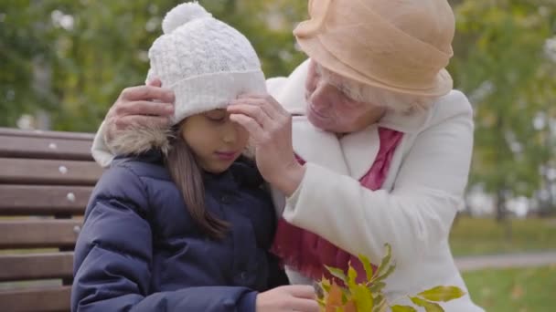 Mulher caucasiana sênior em chapéu de cloche bege e casaco branco fixando o chapéu branco de sua neta. Feliz avó sentada com a jovem menina bonita no banco no parque de outono . — Vídeo de Stock