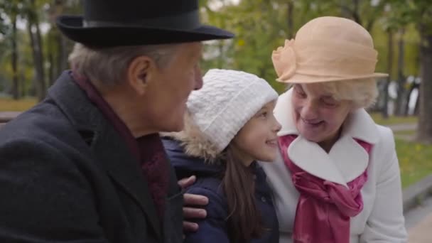 Nahaufnahme eines reifen kaukasischen Paares, das mit seiner Enkelin auf der Bank sitzt und spricht. Großeltern in eleganter klassischer Kleidung verbringen Zeit mit einem fröhlichen jungen Mädchen im Freien. — Stockvideo