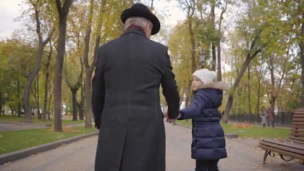 Widok z tyłu dojrzałego białego mężczyzny w eleganckim czarnym kapeluszu i płaszczu spacerującego z wnuczką wzdłuż alejki w jesiennym parku. Całkiem wesołe dziecko ucieka przed dziadkiem.. — Wideo stockowe