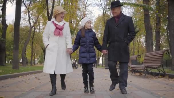 Ein nettes kaukasisches Mädchen mit weißem Hut schlendert mit ihren Großeltern und rennt vorwärts. positives reifes Paar verbringt Freizeit mit ihrer entzückenden Enkelin. — Stockvideo