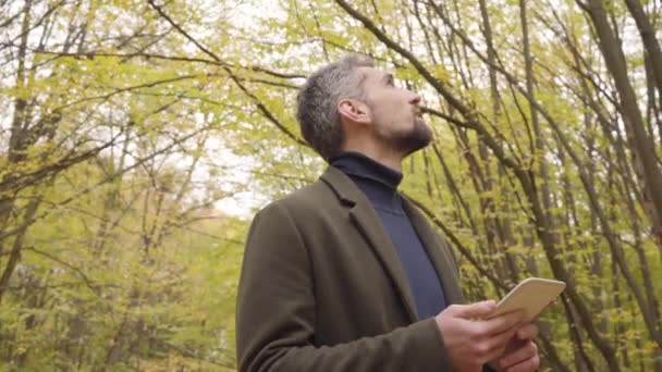 Snygg vit man som tittar upp och runt i skogen och håller i en liten tablett. Självsäker turist njuter av lugn höstdag utomhus. — Stockvideo