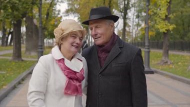 Sonbahar parkında dikilip konuşan zarif elbiseli, olgun beyaz bir çift. Karısı sevimli kocasını yanağından öpüyor ve gülümsüyor..