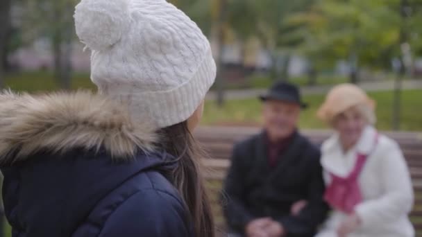 Усміхнена кавказька дівчина у білому капелюсі звертається до бабусі та дідуся, які сидять на лавці на задньому плані і махають руками. Весела дитина проводить осінній день із сім "єю надворі.. — стокове відео