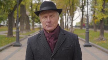 Siyah şapkalı ve ceketli, fındık rengi gözlü, yaşlı, beyaz bir adamın kameraya baktığı yakın çekim. Sonbahar parkında duran olgun bir adam..