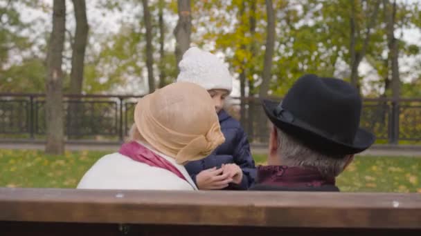 Rückansicht eines älteren kaukasischen Paares, das auf der Bank sitzt und einem fröhlich lächelnden Mädchen zuhört, das vor ihnen springt. Enkelin unterhält ihre Großeltern im Herbstpark. — Stockvideo
