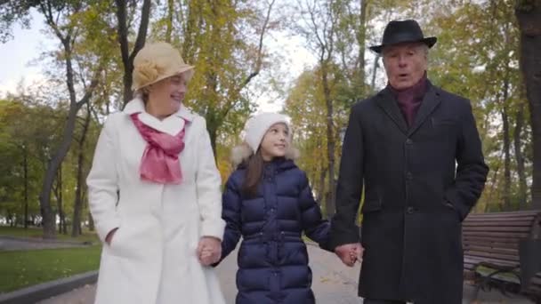 Mooi blank meisje met witte hoed en blauwe jas, wandelend met haar grootouders langs de steeg in het park. Oudere echtpaar gekleed in elegante kleren hand in hand van hun schattige kleindochter. — Stockvideo