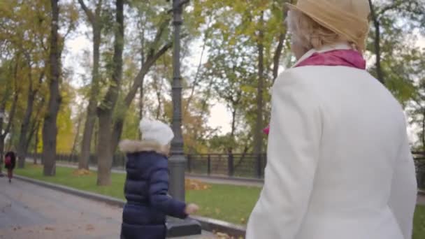 Linda chica caucásica en sombrero blanco y abrigo azul cálido pasar el día de otoño con su abuela en el parque. Mujer madura en sombrero de cloche beige y abrigo blanco hablando con su nieta y sonriendo . — Vídeo de stock