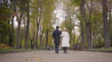 Sonbahar parkında yürüyen ve konuşan olgun, beyaz bir çiftin arka görüntüsü. Siyah elbiseli olgun bir adam ve sokakta dolaşan beyaz ceketli karısı..