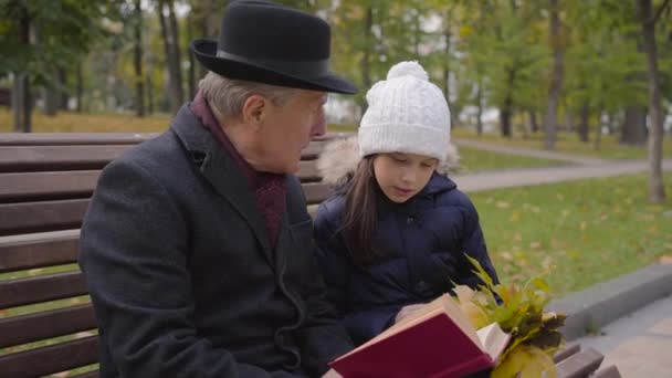 Mature Caucasian man in classic clothes sitting on the bench with his granddaughter and reading a book in red cover. Pretty smiling girl listening to her wise grandfather attentively. — ストック動画