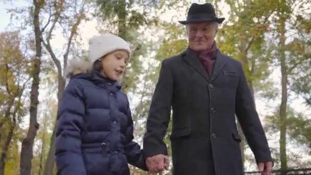 Pretty Caucasian girl in white warm hat spending weekends with her grandfather. Child jumping around senior smiling man in elegant black hat and coat. — ストック動画