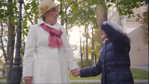 Gammal vit kvinna i beige cloche hatt och vit rock står med sin glada barnbarn på gården av huset. Liten leende flicka hoppar upp och lyssnar på sin mormor. — Stockvideo