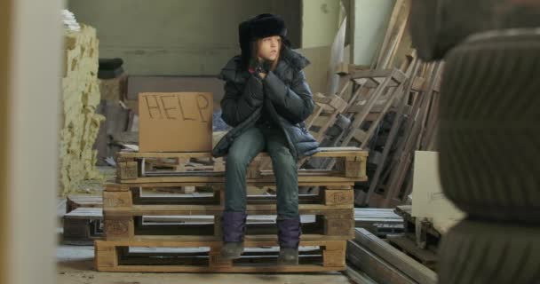 Pequeno refugiado sírio em roupas quentes sujas sentado em paletes no canteiro de obras com cartão Help. Menino solitário sem-teto esperando por uma esmola . — Vídeo de Stock