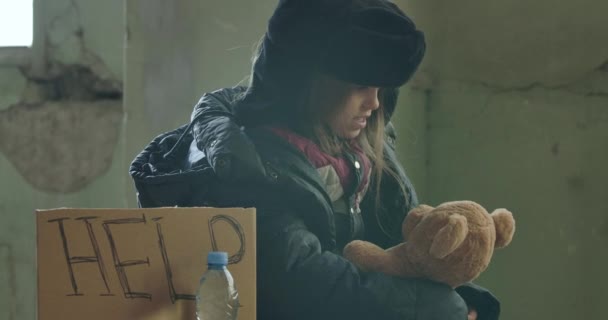 Ritratto di una rifugiata siriana in abiti sporchi seduta con Aiuto cartone e parlando con il suo orsacchiotto. Bambino senzatetto solitario che indossa cappello con paraorecchie in attesa di qualche aiuto . — Video Stock
