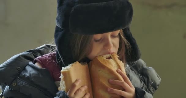 Retrato de cerca de una chica sin hogar con sombrero con orejeras comiendo ávidamente la barra de pan. Refugiado hambriento viviendo en las calles . — Vídeos de Stock