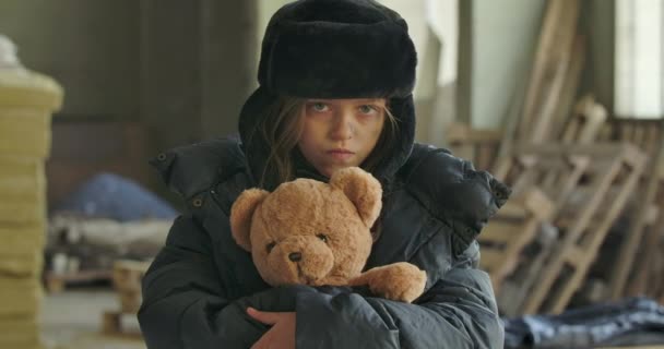 Portrait of a homeless girl with grey eyes wearing hat with earflaps looking at the camera and hugging mudtard teddy bear. Hopeless refugee living on the streets. — Stock Video