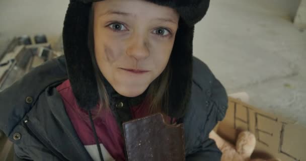 Emocionada chica caucásica sin hogar con la cara sucia y los ojos grises codiciosamente comiendo la barra de chocolate. Pequeño refugiado en sombrero con orejeras teniendo comida para comer . — Vídeos de Stock