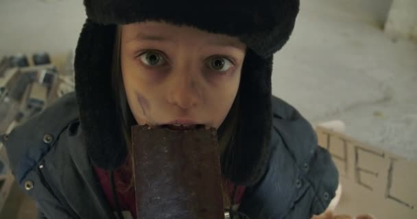 Skrämd vit hemlös tjej med smutsigt ansikte och grå ögon som glupskt äter chokladkakan. Liten flykting i hatt med öronlappar har mat att äta. — Stockvideo