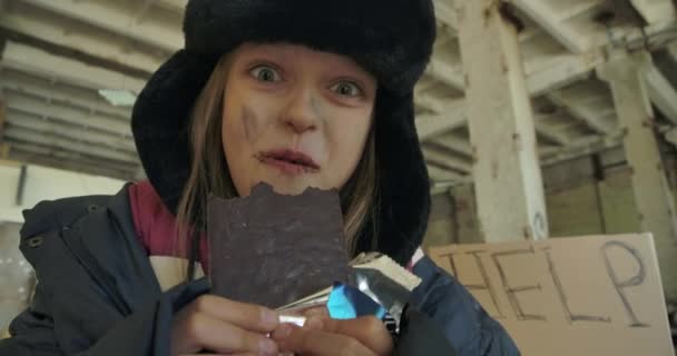 Retrato de close-up de uma menina sem-teto caucasiana com rosto sujo e olhos grisalhos comendo avidamente a barra de chocolate. Criança excitada de chapéu com protetores auriculares que têm comida para comer . — Vídeo de Stock