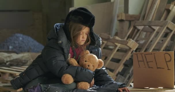 Chica caucásica somnolienta en ropa de invierno sentado con el oso de peluche en el sitio de construcción. Niño enfermo y agotado que vive en las calles . — Vídeos de Stock
