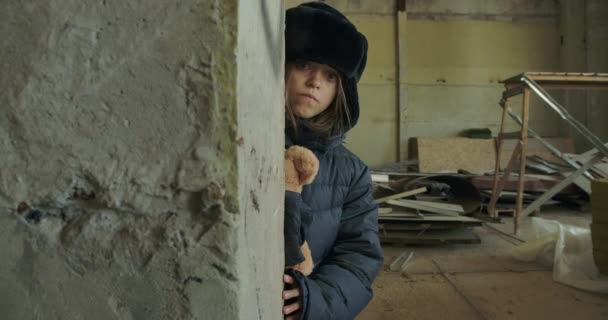 Retrato de menina sem-teto caucasiana em roupas de inverno escondido no local de construção com o urso de pelúcia. Criança assustada andando por trás da parede e olhando para a câmera . — Vídeo de Stock