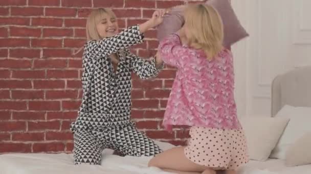 Two blonde Caucasian friends fighting with pillows at the white bed and laughing. Beautiful women having pajama party in the bedroom at home.