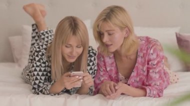 Beyaz yumuşak yatağa uzanmış beyaz pijamalı iki beyaz sarışın kız akıllı telefona bakıyor. Mutlu, gülümseyen kadınlar evde pijama partisi veriyorlar..