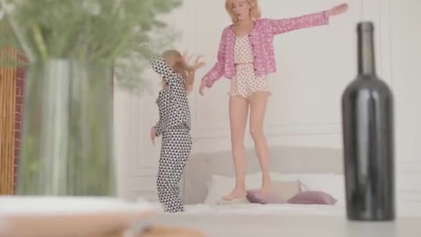 Две молодые блондинки прыгают на мягкую кровать и смеются. Весёлые друзья устраивают пижамную вечеринку дома. Сосредоточьтесь на бутылке вина, стоящей на переднем плане . — стоковое видео