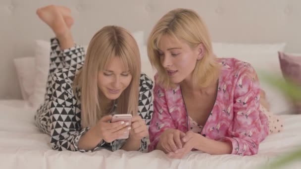 Duas jovens loiras caucasianas de pijama deitadas na cama macia branca e olhando para o smartphone. Feliz sorrindo mulheres fazendo festa de pijama em casa . — Vídeo de Stock