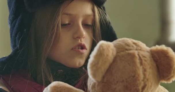 Portrait of a cute homeless girl in hat with earflaps talking to the beige teddy bear. Desperate refugee waiting for a help. — Stock Video