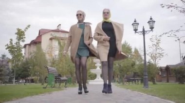 Şehir parkında yürüyen bej ceketli, kısa elbiseli ve güneş gözlüklü iki zarif olgun Kafkas bayan arkadaş. Büyüleyici yetişkin kadınlar sonbahar günlerini dışarıda geçirirler..