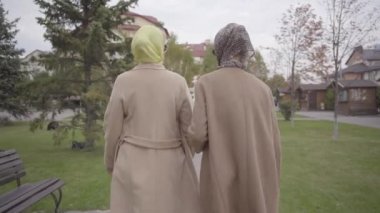 Beyaz önlüklü, türbanlı ve güneş gözlüklü iki güzel beyaz kadının şehir parkında yürüyüşü. Büyüleyici yetişkin bayan arkadaşlar birlikte seyahat ediyorlar..