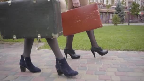 Vista lateral de las piernas en tacones altos de dos mujeres sosteniendo viejas bolsas de viaje y caminando. Elegantes amigas viajando juntas . — Vídeos de Stock