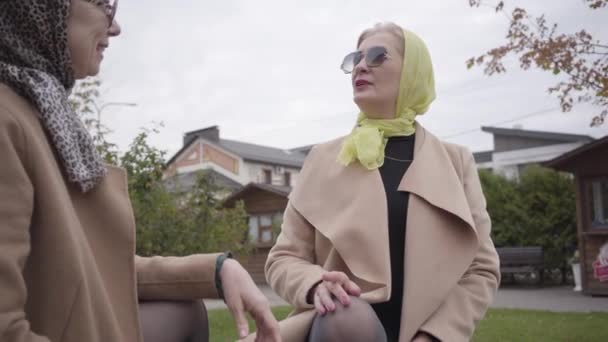 Gros plan de deux femmes blanches adultes portant un foulard et des lunettes de soleil assises sur le banc et parlant. Élégantes amies mûres passent du temps ensemble à l'extérieur . — Video