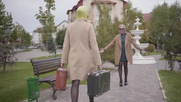 Dvě dospělé kavkazské kamarádky se během cesty setkávají. Elegantní žena se dvěma cestovními taškami kráčející k usmívající se kamarádce a objímající ji. — Stock video