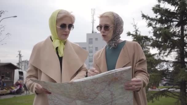 Дві зрілі жінки в хустках і сонцезахисних окулярах тримають карту і розмовляють. Старші леді подорожують після виходу на пенсію разом . — стокове відео