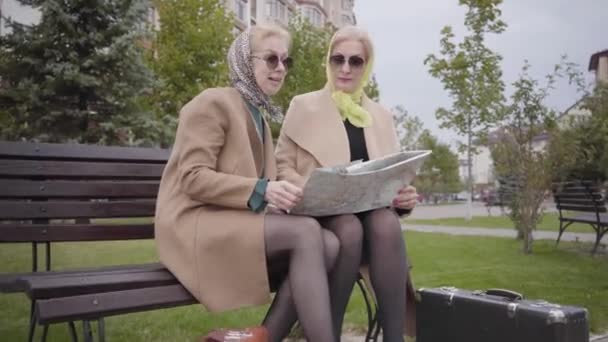 Senior amigas caucásicas en pañuelos para la cabeza y gafas de sol sentadas en el banco y examinando el mapa. Maduras señoras elegantes viajando después de la jubilación juntos . — Vídeos de Stock