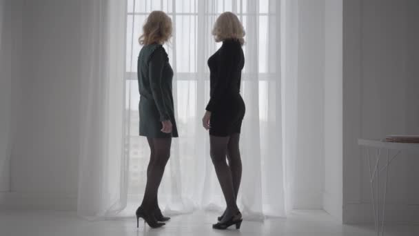 Två mogna blonda vita kvinnor i korta eleganta klänningar som står bredvid fönstret och pratar. Rika seniora affärsslavar diskuterar semestrar. — Stockvideo