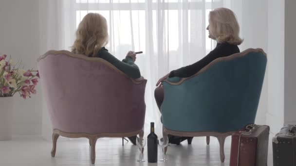 Rückansicht reifer blonder kaukasischer Frauen, die in bequemen Sesseln sitzen und miteinander reden. Seniorinnen verbringen Urlaub mit einer Flasche Wein und Zigarren. — Stockvideo